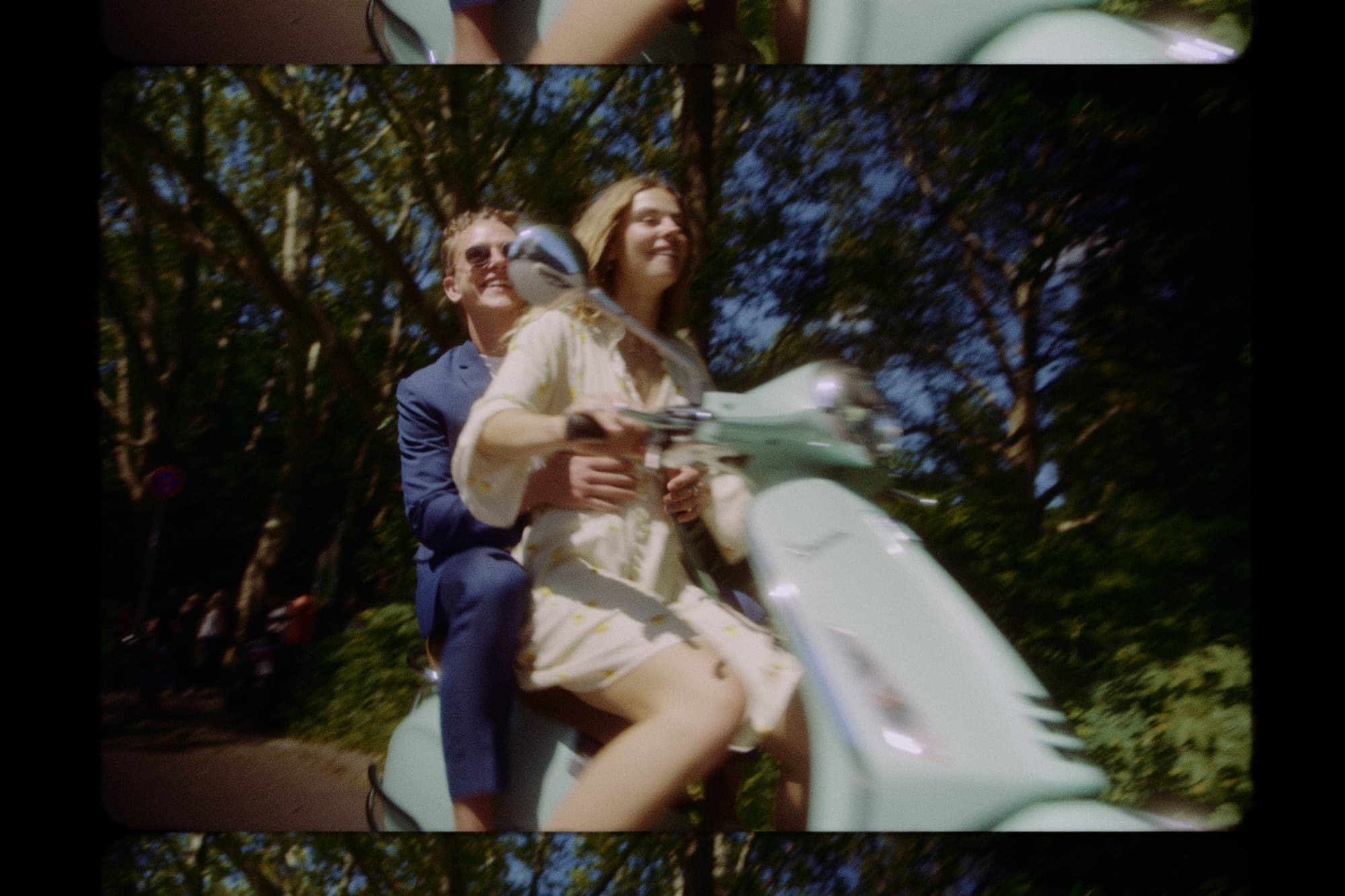 Super 8 style still of a couple riding a scooter through trees on their wedding day